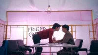 Dois homens com rostos próximos sobre uma mesa. Um quarto rosa iluminado com letras FRIEND CR no fundo. Clima intenso.