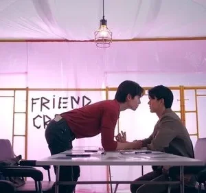 Dois homens com rostos próximos sobre uma mesa. Um quarto rosa iluminado com letras FRIEND CR no fundo. Clima intenso.