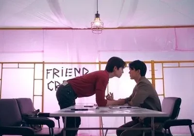 Dois homens com rostos próximos sobre uma mesa. Um quarto rosa iluminado com letras FRIEND CR no fundo. Clima intenso.