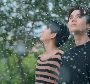 Dois jovens olham para cima enquanto flocos brancos caem suavemente. Ambiente exterior verde e calmo.