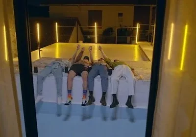 Quatro jovens deitados num terraço à noite, com as mãos para cima. Luzes neon amarelas iluminam o ambiente urbano e descontraído.