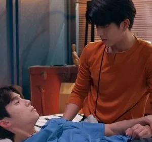 Dois jovens, um deitado e um de laranja em pé, trocam olhares enquanto um segura o braço do outro. Cena íntima em ambiente interno.