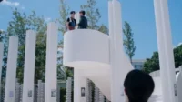 Estrutura arquitetônica branca e moderna com pilares. Dois homens conversam no topo, sob céu azul claro. Um terceiro observa de baixo.
