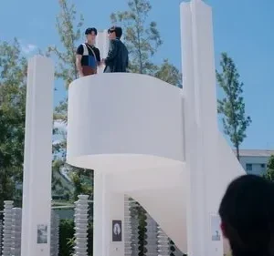 Estrutura arquitetônica branca e moderna com pilares. Dois homens conversam no topo, sob céu azul claro. Um terceiro observa de baixo.