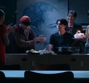 Grupo de 6 jovens celebra um aniversário em ambiente escuro. Um segura bolo com velas, outros batem palmas e sorriem. Clima de festa.