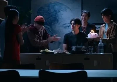 Grupo de 6 jovens celebra um aniversário em ambiente escuro. Um segura bolo com velas, outros batem palmas e sorriem. Clima de festa.