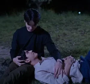 Dois homens em um campo escuro. Um sentado olha carinhosamente para o outro deitado com a cabeça em seu colo. Mãos próximas, clima íntimo.