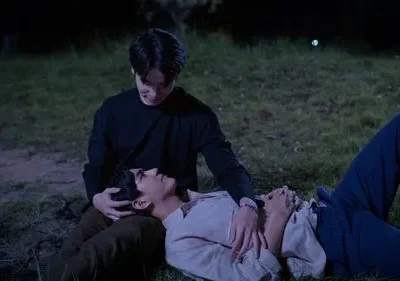 Dois homens em um campo escuro. Um sentado olha carinhosamente para o outro deitado com a cabeça em seu colo. Mãos próximas, clima íntimo.