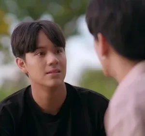 Jovem de cabelo escuro e camiseta preta olha pensativo para outro homem de costas, em fundo verde desfocado.