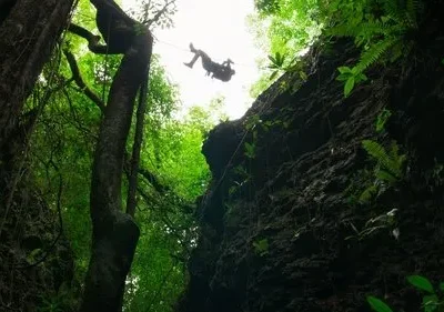 Silhueta de pessoa em rapel numa fenda na selva. Paredes rochosas e árvores. Folhagem verde e céu claro criam um clima aventureiro.