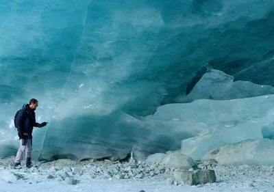 Homem em roupa de inverno explora uma caverna de gelo azul-turquesa. Ele toca a parede gelada num ambiente rochoso e frio.
