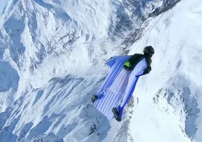 Pessoa em wingsuit azul voando sobre vasta paisagem de montanhas nevadas. Cena de ação com clima de aventura extrema.