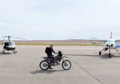 Pessoa em moto de trilha numa pista de pouso, entre um helicóptero e um avião, com deserto ao fundo. Cenário de aventura.