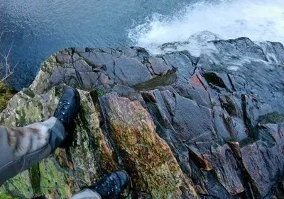 Perspectiva de primeira pessoa: botas de trilha na beira de um precipício rochoso, com água furiosa abaixo.