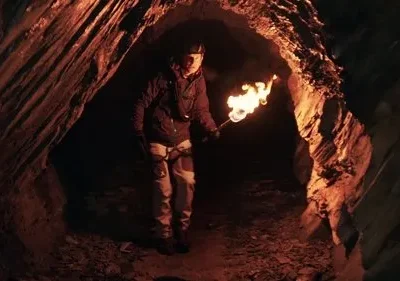 Uma pessoa com capacete e tocha explora uma caverna escura. A luz laranja destaca as paredes rochosas, criando um clima misterioso.