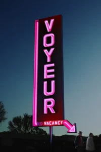 Letreiro neon rosa VOYEUR com NO VACANCY se destaca contra um céu azul-escuro. Dois homens sombrios estão em primeiro plano. Clima enigmático.