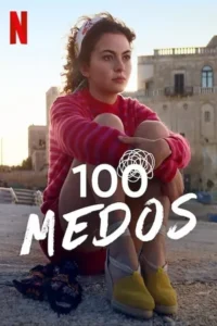 Mulher jovem sentada,abraçando os joelhos,olha pensativa em cenário costeiro com edifícios antigos. Céu claro e clima calmo.