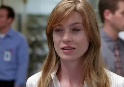 Mulher de cabelo loiro-claro e olhar pensativo em foco. Homens borrados com crachás ao fundo, em ambiente profissional.