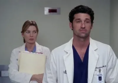 Um médico sério de jaleco no primeiro plano olha para a frente. Uma médica com pastas, de expressão preocupada, está atrás dele no hospital.