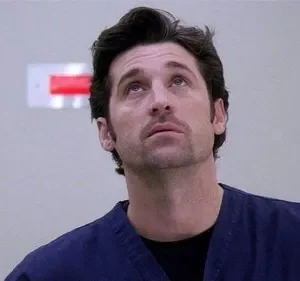 Homem de scrubs azuis e barba rala olhando para cima com expressão pensativa. Ambiente claro, com um sinal vermelho na parede.