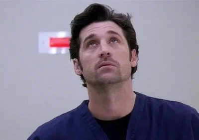 Homem de scrubs azuis e barba rala olhando para cima com expressão pensativa. Ambiente claro, com um sinal vermelho na parede.