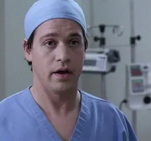 Um médico jovem de scrubs azuis e touca, com boca aberta e olhar preocupado, num hospital com aparelhos.