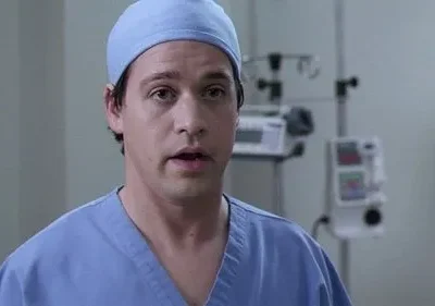 Um médico jovem de scrubs azuis e touca, com boca aberta e olhar preocupado, num hospital com aparelhos.