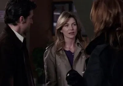 Uma mulher de casaco bege conversa com outra de costas, enquanto um homem de casaco escuro as observa. Ambiente interno.