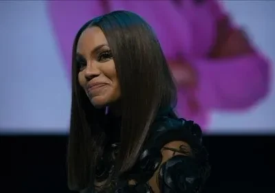 Mulher com cabelo escuro liso e sorriso sutil. Veste top preto texturizado. Fundo com projeção desfocada em tons de magenta e azul.