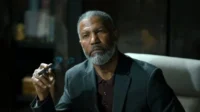 Homem negro de barba grisalha e expressão séria segura charuto fumegante. Fumaça azul sobe em ambiente escuro e elegante.