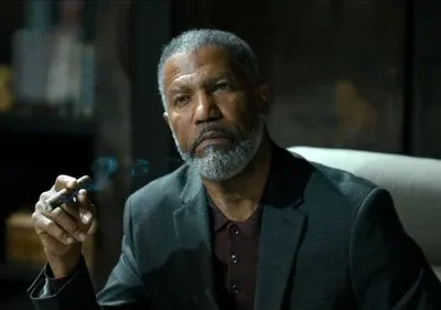 Homem negro de barba grisalha e expressão séria segura charuto fumegante. Fumaça azul sobe em ambiente escuro e elegante.