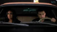 Uma mulher e um homem jovens no carro à noite. Ambos olham sérios para frente, com luzes urbanas borradas no fundo. Sugere drama.