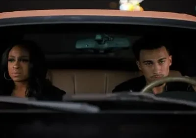 Uma mulher e um homem jovens no carro à noite. Ambos olham sérios para frente, com luzes urbanas borradas no fundo. Sugere drama.