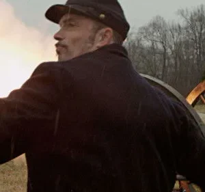 Homem de uniforme militar e bigode observa. Canhão fumegante e outro soldado no campo. Clima de batalha histórica.