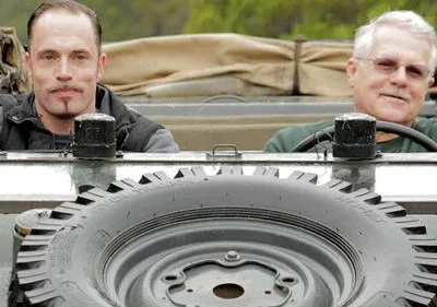 Dois homens, um jovem sério e um idoso sorridente, em veículo robusto com pneu sobressalente. Clima de aventura.