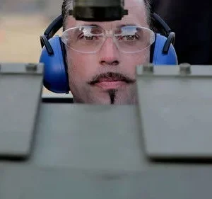 Rosto de homem com bigode e cavanhaque, óculos e fones azuis, focado. Ele espia por trás de uma estrutura metálica. Clima de tensão.