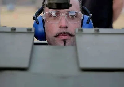 Rosto de homem com bigode e cavanhaque, óculos e fones azuis, focado. Ele espia por trás de uma estrutura metálica. Clima de tensão.