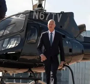 Homem de terno, com pasta verde, caminha após descer de helicóptero preto. Outro sobe. Cenário urbano, clima de negócios.