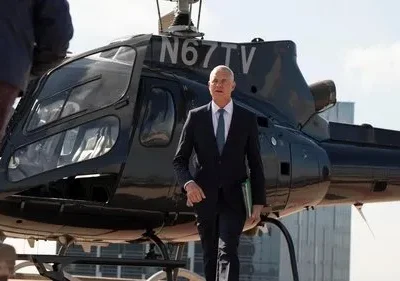 Homem de terno, com pasta verde, caminha após descer de helicóptero preto. Outro sobe. Cenário urbano, clima de negócios.