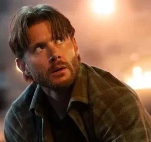 Homem barbudo em camisa xadrez olha tenso para o alto. Fundo escuro com chamas e fumaça, indicando perigo.