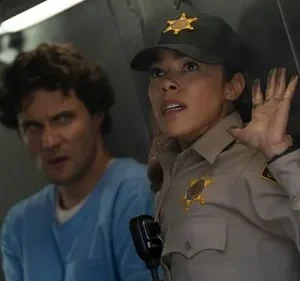 Uma xerife assustada com mãos sujas e um homem de camisa azul com olhar intenso em um cenário tenso.