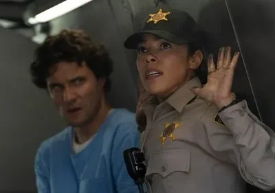 Uma xerife assustada com mãos sujas e um homem de camisa azul com olhar intenso em um cenário tenso.