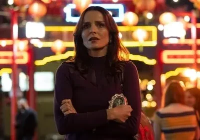 Mulher com expressão preocupada segura foto, em rua noturna iluminada por lanternas e luzes de neon. Ambiente vibrante, clima tenso.