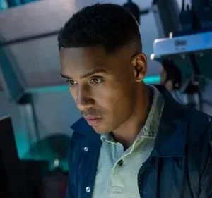 Jovem negro de jaqueta azul olha focado para monitor, em ambiente escuro com brilhos azuis. Clima de tensão.