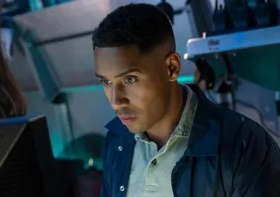 Jovem negro de jaqueta azul olha focado para monitor, em ambiente escuro com brilhos azuis. Clima de tensão.