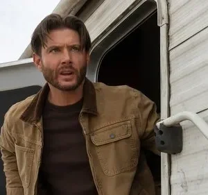 Homem barbudo com jaqueta marrom sai de um trailer branco, olhando surpreso e tenso para algo fora da tela. Céu claro.