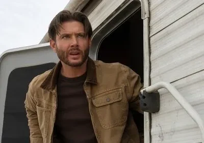 Homem barbudo com jaqueta marrom sai de um trailer branco, olhando surpreso e tenso para algo fora da tela. Céu claro.