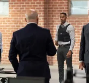 Quatro pessoas em ambiente de tijolos. Mulher e jovem com coletes táticos, dois homens de terno. Atmosfera séria e profissional.