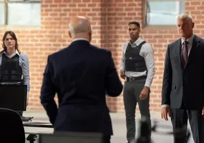 Quatro pessoas em ambiente de tijolos. Mulher e jovem com coletes táticos, dois homens de terno. Atmosfera séria e profissional.