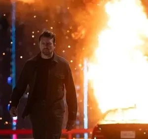 Homem de casaco escuro caminha à noite. Um carro em chamas está à direita, luzes azuis verticais à esquerda. Clima tenso de ação.